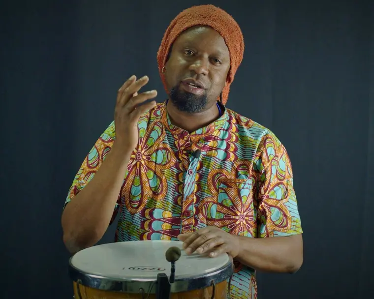 Master Adson Junior, in an orange cap and African-print shirt, teaching behind a drum.